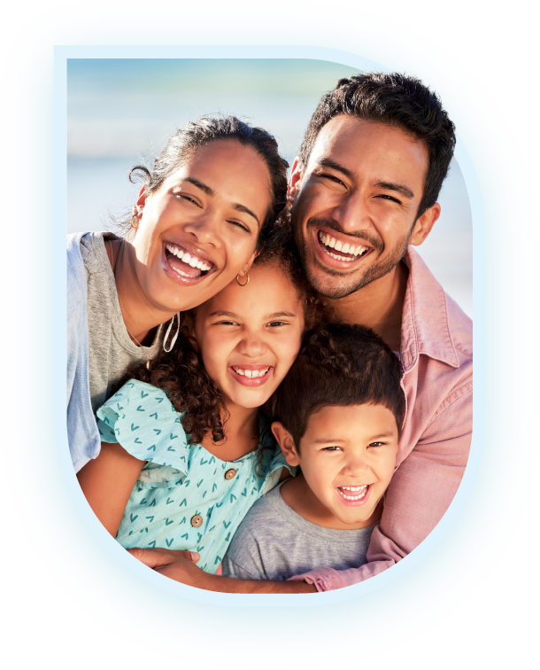 A happy family, consisting of a mother, father, daughter, and son, hugging and smiling together against a beach backdrop| Pleasant Dental